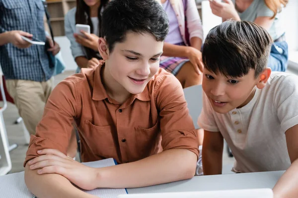 Улыбающийся школьник смотрит на размытый цифровой планшет рядом с азиатским другом — стоковое фото
