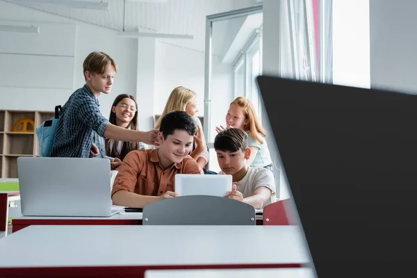 Interracial schoolkids using digital tablet near laptops and friends — Stock Photo