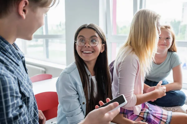 Lächelnde Schülerin blickt verschwommene Mitschülerin mit Smartphone im Klassenzimmer an — Stockfoto