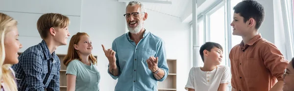 Mature teacher pointing with fingers near interracial smiling kids in classroom, banner — Stock Photo