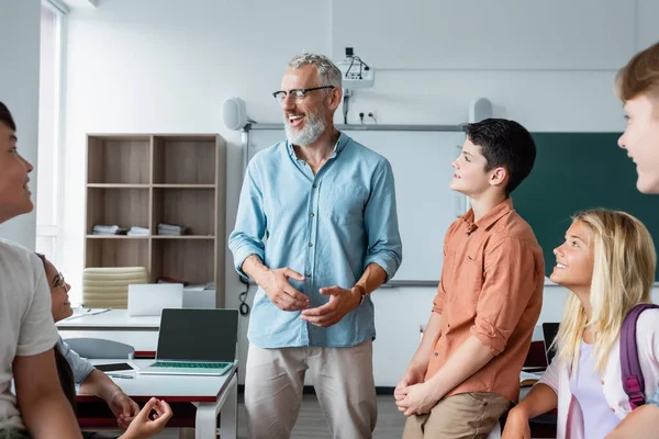 Fröhlicher Lehrer im Gespräch mit Schülern im unscharfen Vordergrund im Klassenzimmer — Stockfoto