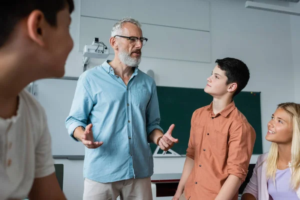 Grauhaarige Lehrerin gestikuliert im Gespräch mit Teenagern im Klassenzimmer — Stockfoto