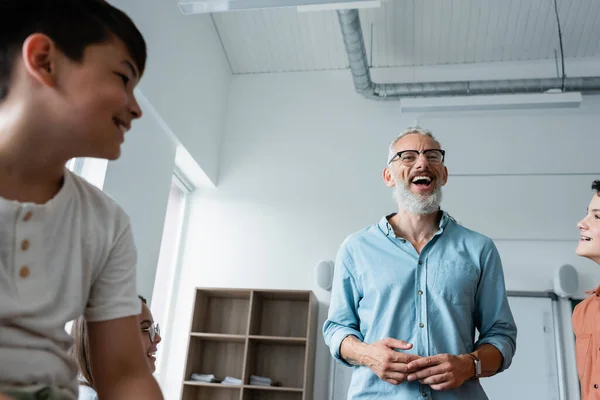 Low angle view of bearded teacher laughing near multiethnic pupils in classroom — Stock Photo