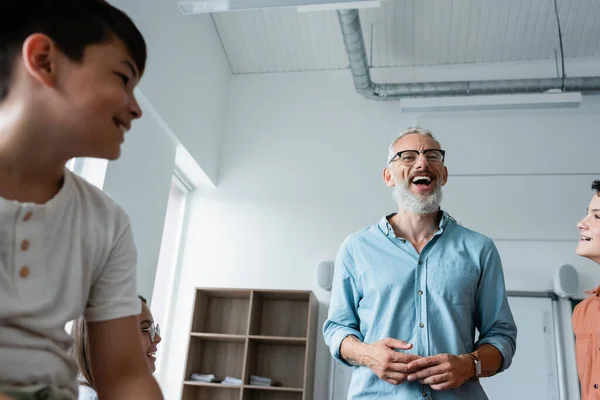 Vista ad angolo basso di insegnante barbuto ridere vicino a alunni multietnici in classe — Foto stock