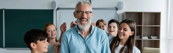 Excited middle aged teacher laughing with closed eyes near happy multicultural pupils, banner — Stock Photo