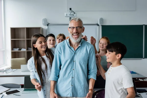 Colegas multiétnicos alegres perto de professor feliz rindo com os olhos fechados em sala de aula — Fotografia de Stock