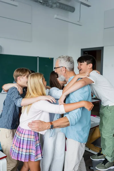 Adolescents multiculturels embrassant professeur aux cheveux gris dans la salle de classe — Photo de stock