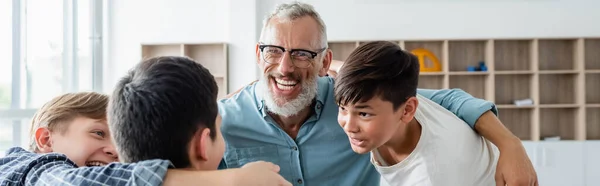 Multicultural boys and cheerful middle aged teacher embracing in classroom, banner — Stock Photo