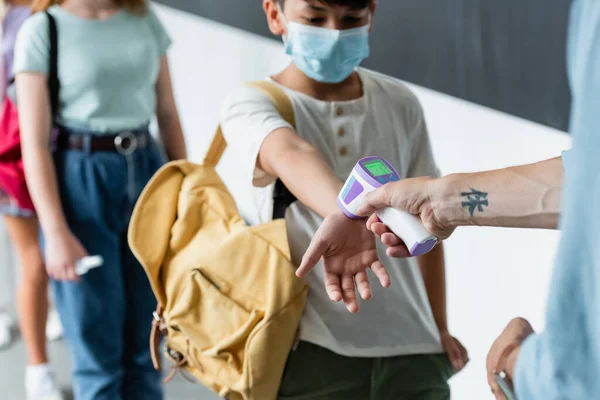 Cropped view of teacher measuring temperature of blurred asian boy in protective mask with pyrometer — Stock Photo