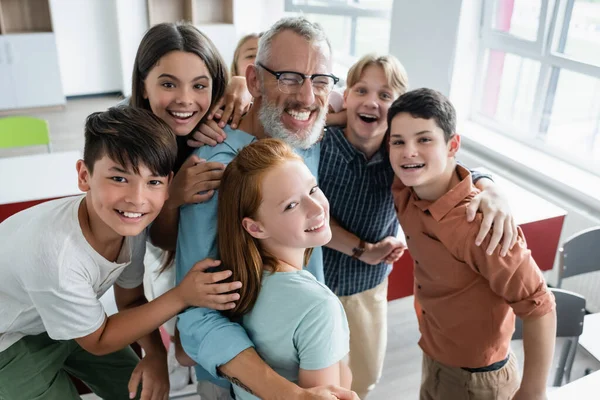 Fröhliche multiethnische Klassenkameraden, die in die Kamera schauen, während sie fröhliche Lehrer umarmen, die mit geschlossenen Augen lachen — Stockfoto
