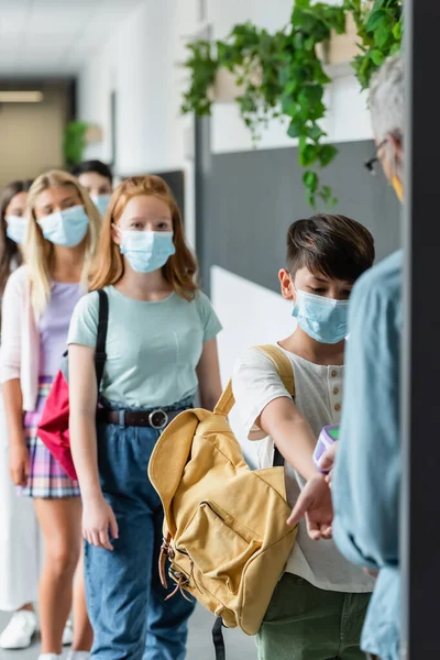 Blurred teacher checking temperature of multiethnic pupils in medical masks with pyrometer — Stock Photo