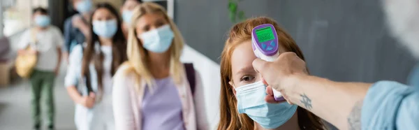 Pirómetro en la mano del profesor que mide la temperatura de la colegiala en máscara médica, pancarta - foto de stock