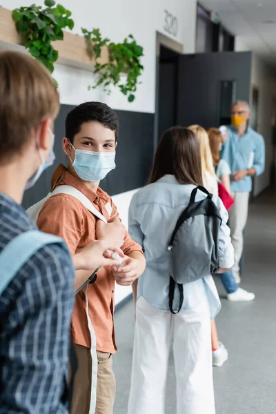 Niño en máscara médica tomando desinfectante de manos de un amigo mientras está de pie en la fila en el pasillo de la escuela - foto de stock