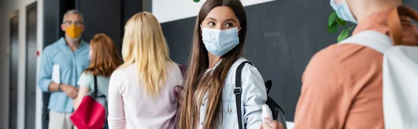 Adolescente en máscara médica tomando desinfectante de manos de amigo cerca de compañeros de clase y profesor borroso, pancarta - foto de stock