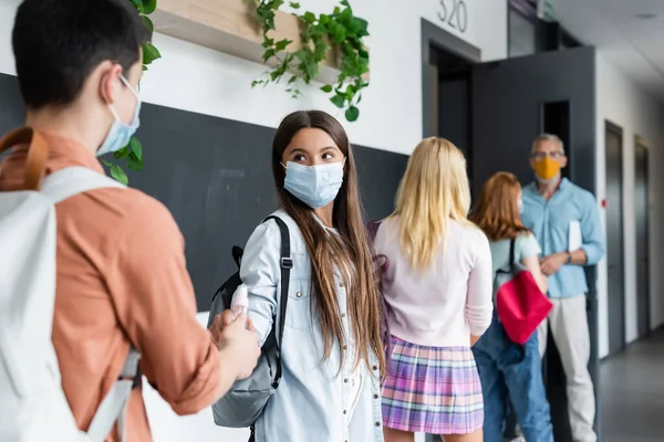 Teenage boy giving hand antiseptic to girl in medical mask near blurred classmates and teacher — Stock Photo