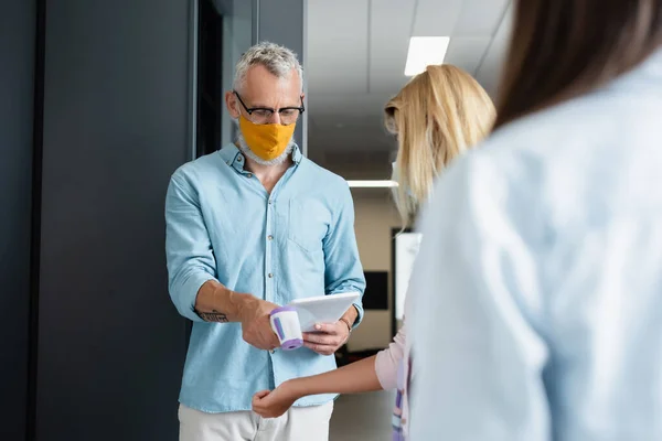 Teacher in orange medical mask checking temperature of teenage girl in school — Stock Photo