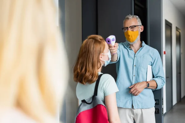 Middle aged teacher in orange medical mask measuring temperature of schoolgirl with pyrometer — Stock Photo