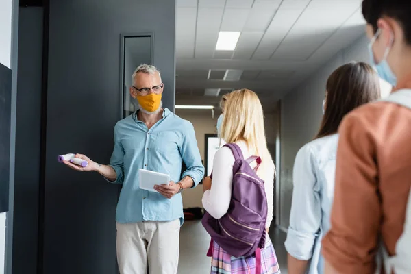 Reife Lehrerin mit Pyrometer und digitalem Tablet lädt Schülerin in medizinischer Maske ins Klassenzimmer ein — Stockfoto