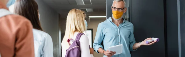 Maestro en máscara médica sosteniendo pirómetro mientras señala en el aula cerca de colegiala, pancarta - foto de stock