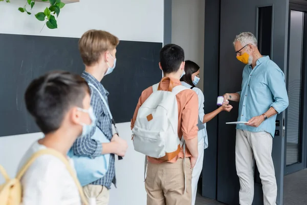 Mature teacher in medical mask checking temperature of teenage pupils with pyrometer — Stock Photo