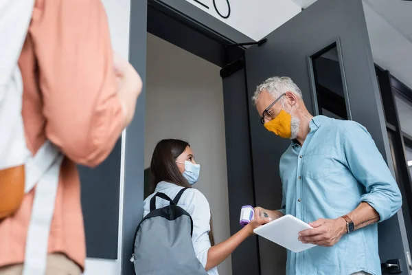 Low angle view of teacher in orange medical mask checking temperature of schoolgirl with pyrometer — Stock Photo