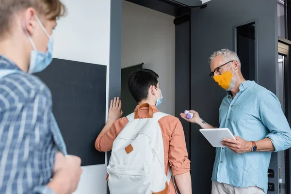 Middle aged teacher with pyrometer and digital tablet inviting schoolboy in medical mask into classroom — Stock Photo