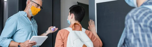 Вчителька з пірометром і цифровим планшетом запрошує школяра в медичну маску в клас, банер — стокове фото
