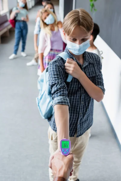 Teenage boy in medical mask near teacher measuring his temperature with pyrometer — Stock Photo