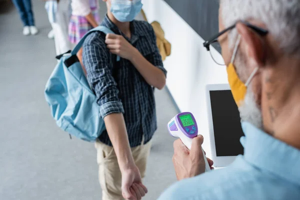 Ausgeschnittene Ansicht von Schülern in medizinischen Masken in der Nähe verschwommener Lehrer mit digitalem Tablet und Pyrometer — Stockfoto