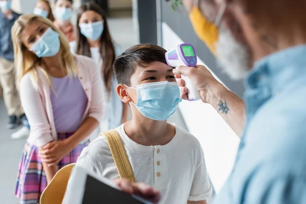 Borrosa profesora medir temperatura de asiático chico con pyrometer cerca borrosa pupilas en médico máscaras - foto de stock