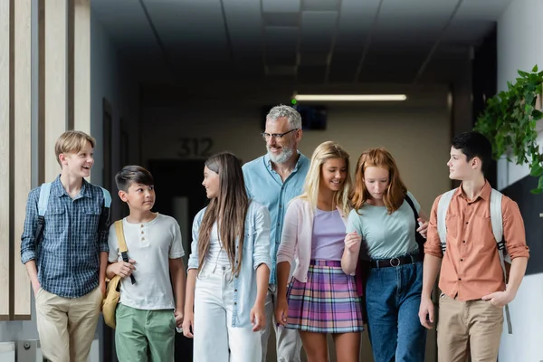 Adolescentes multiétnicos caminando con un profesor sonriente en el pasillo de la escuela - foto de stock