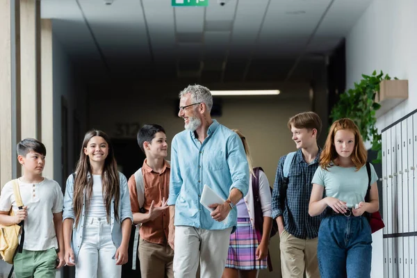 Adolescente hablando con un profesor sonriente mientras camina con compañeros de clase multiétnicos en el salón de clases - foto de stock