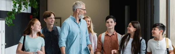 Happy multicultural teenagers looking at middle aged teacher while walking in school corridor, banner — Stock Photo