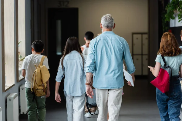 Back view of teacher with digital tablet and pupils with backpacks walking along school corridor — Stock Photo