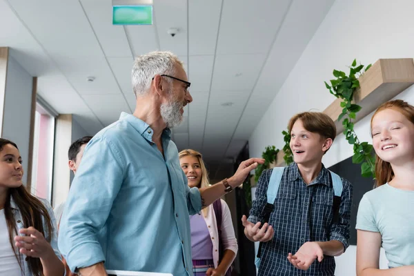 Mature teacher talking to excited boy in school corridor near classmates — Stock Photo