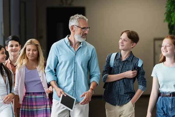 Middle aged teacher with digital tablet walking in school hall with teenage pupils — Stock Photo