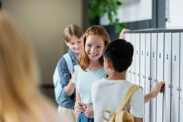 Enfoque selectivo de colegiala pelirroja sonriendo en el pasillo de la escuela cerca de adolescentes borrosos - foto de stock