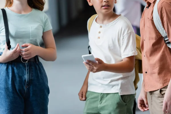 Vista ritagliata dello scolaro con telefono cellulare vicino ai compagni di classe — Foto stock