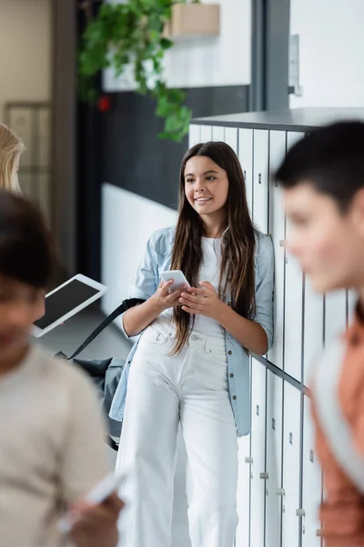 Morena chica con teléfono inteligente sonriendo cerca borrosa compañeros de clase en la sala de la escuela — Stock Photo