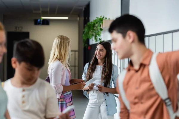 Teenage girls with gadgets talking in school corridor near blurred pupils — Stock Photo