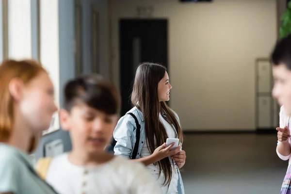 Alegre colegiala sosteniendo el teléfono móvil cerca borrosa compañeros de clase en la sala de la escuela - foto de stock