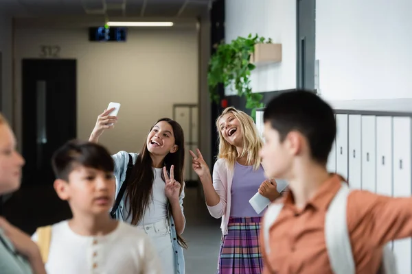 Cheerful girls showing victory gesture while taking selfie near blurred classmates — Stock Photo