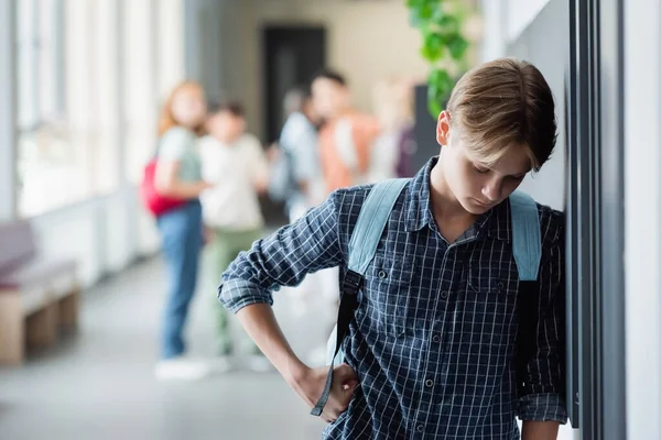 Upset schoolboy standing alone with bowed head near blurred pupils in school corridor — Stock Photo