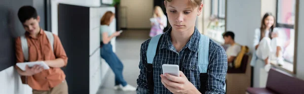 Teenage boy using mobile phone near blurred classmates in school corridor, banner — Stock Photo