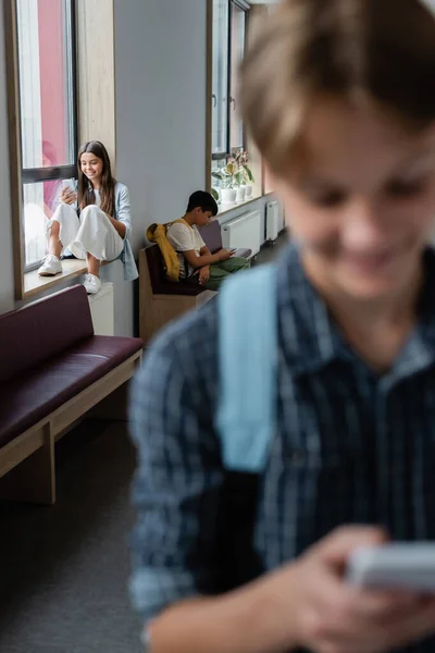 Adolescente offuscata con smartphone vicino ai compagni di classe utilizzando gadget sul davanzale della finestra e panchina in corridoio — Foto stock