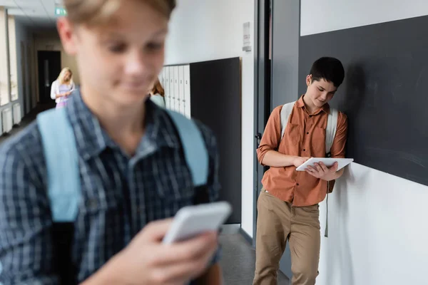 Adolescent garçon en utilisant tablette numérique près de camarade de classe avec téléphone mobile au premier plan flou — Photo de stock