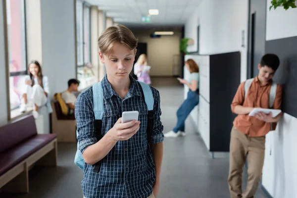 Teenage boy looking at mobile phone near blurred pupils in school corridor — Stock Photo