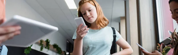 Rousse fille en utilisant smartphone près flous camarades de classe avec des gadgets, bannière — Photo de stock