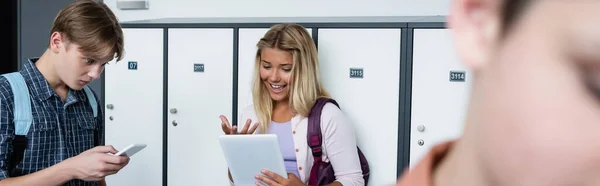 Excitado chica gesto mientras se utiliza la tableta digital cerca de compañero de clase con el teléfono inteligente, bandera - foto de stock