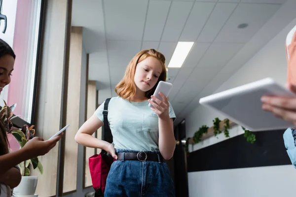 Rothaariges Mädchen schaut mit Gadgets auf Handy in der Nähe verschwommener Teenager — Stockfoto